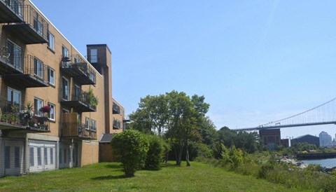A row of apartment buildings with balconies overlook a grassy area and a river.