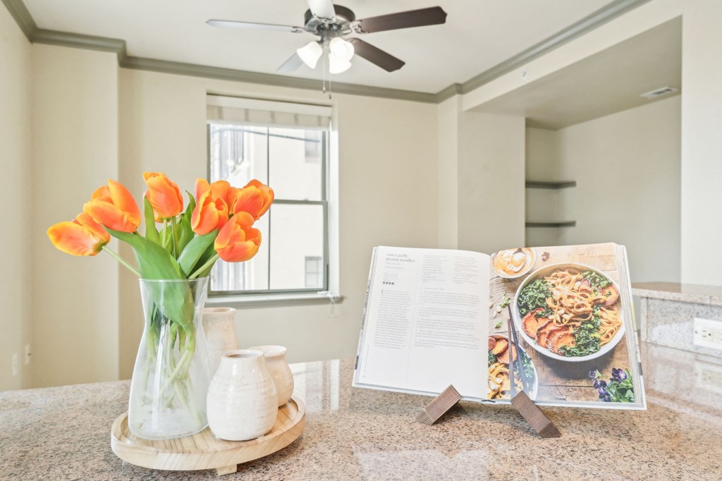 A dining table with a vase of orange flowers, a white jug, and a cookbook open on the page with a recipe.