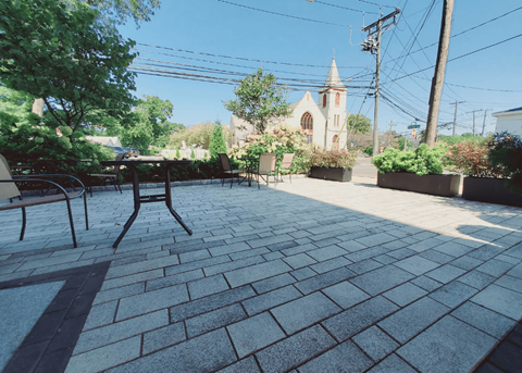 A church with a steeple is in the background of a brick patio with chairs and tables.