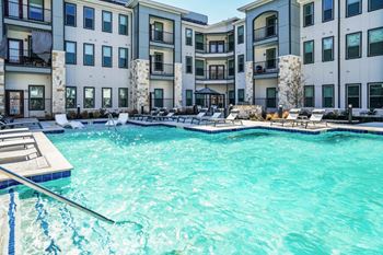 A swimming pool in front of a building with a deck and lounge chairs.