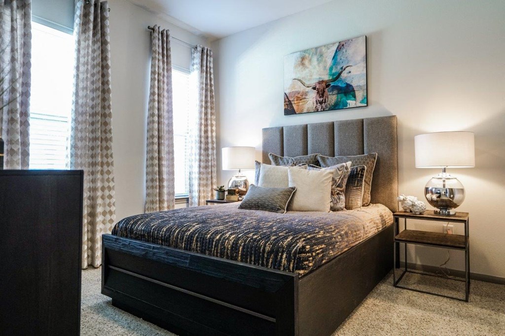 A bedroom with a large bed, a painting on the wall, and a window with curtains at The Grand at Manor Apartments, Manor, TX