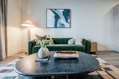 A living room with a green couch, a round table with a plate of food, and a painting on the wall.