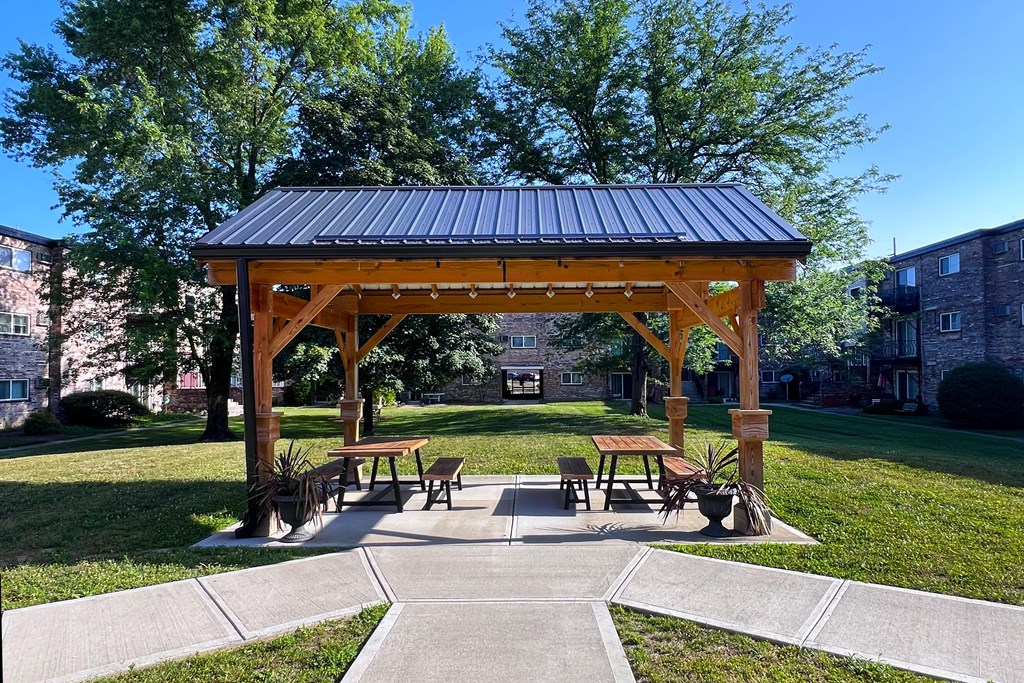 A wooden pavilion with a black roof is surrounded by benches and trees.