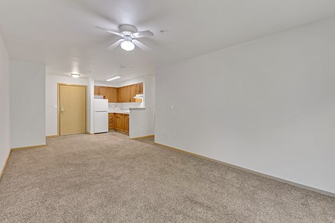 the living room and kitchen of an apartment with carpeting and a ceiling fan