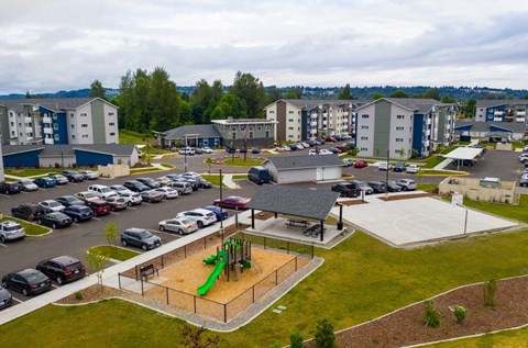 A playground with a green slide is surrounded by a parking lot and apartment buildings.