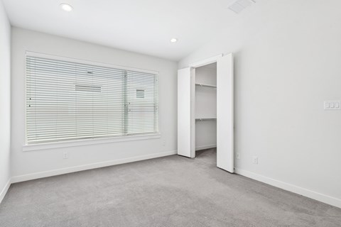 A room with a carpeted floor, a window with blinds, and a door leading to another room.