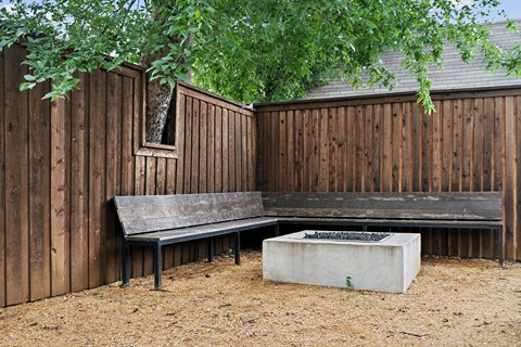 A bench sits in front of a wooden fence with a concrete fire pit in front of it.