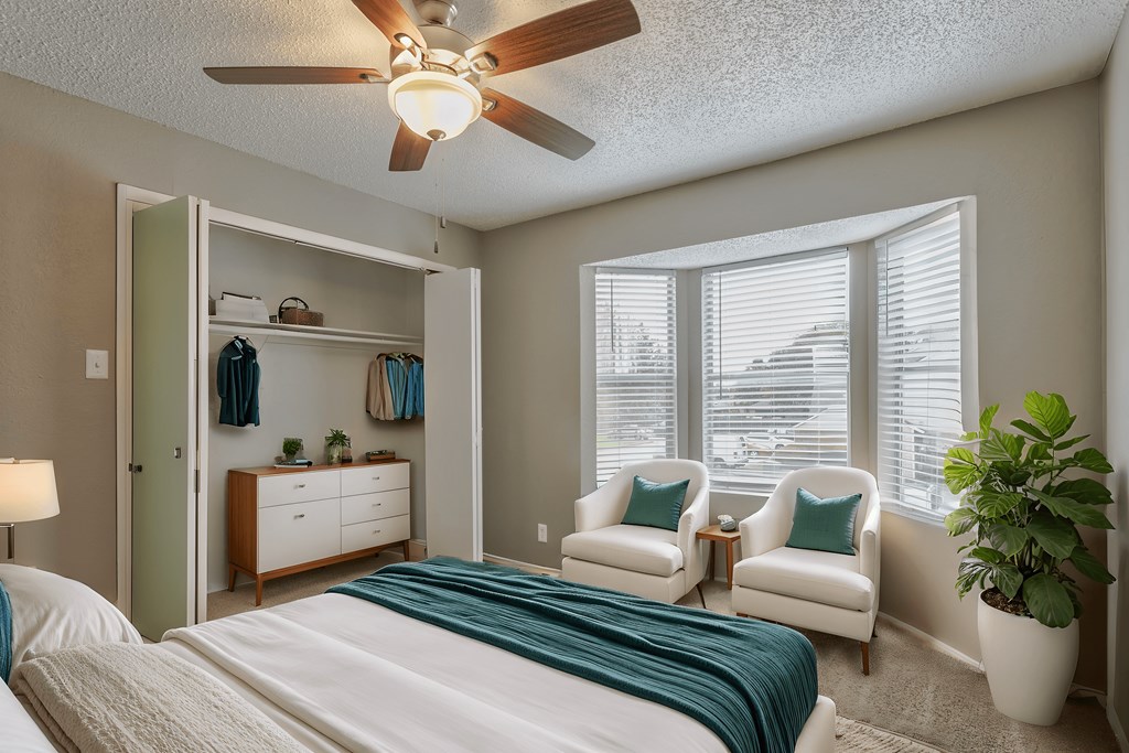 A bedroom with a bed, two chairs, a ceiling fan, and a potted plant.