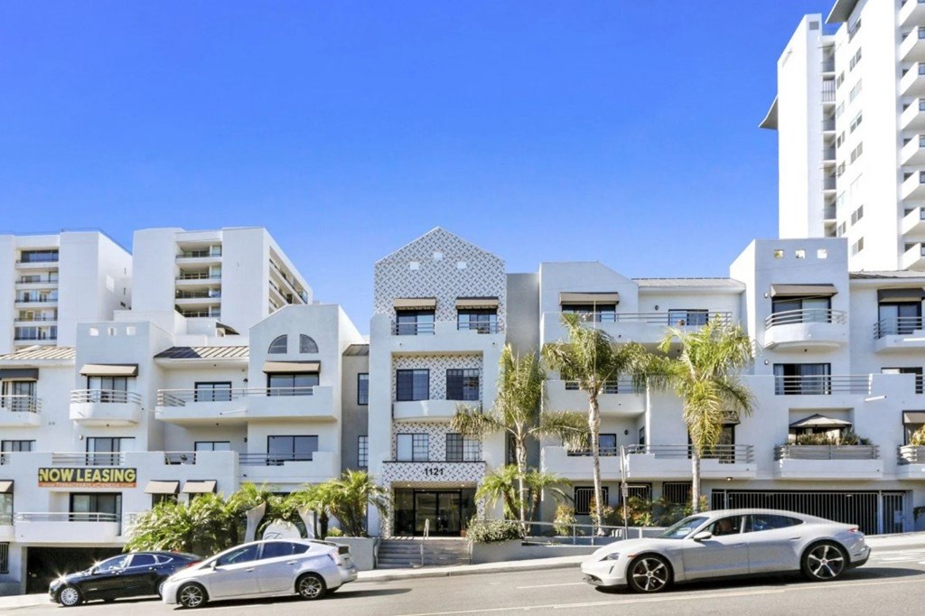 A modern apartment complex with a "Now Leasing" sign is surrounded by palm trees.