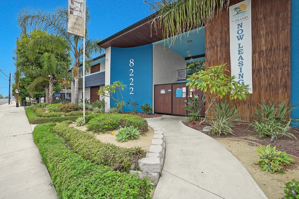 A building with a blue wall and a sign that says "NOW LEASING" is surrounded by greenery.