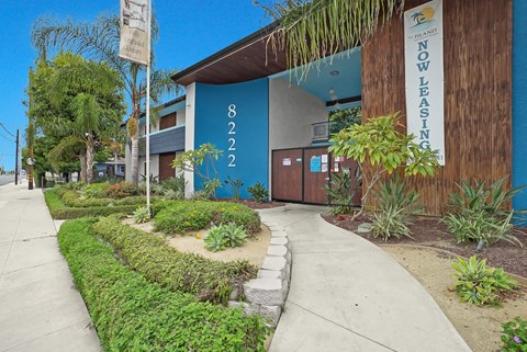 A building with a blue wall and a sign that says "NOW LEASING" is surrounded by greenery.