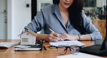 A woman is writing on a notepad with a small model house and a pen in her hand.