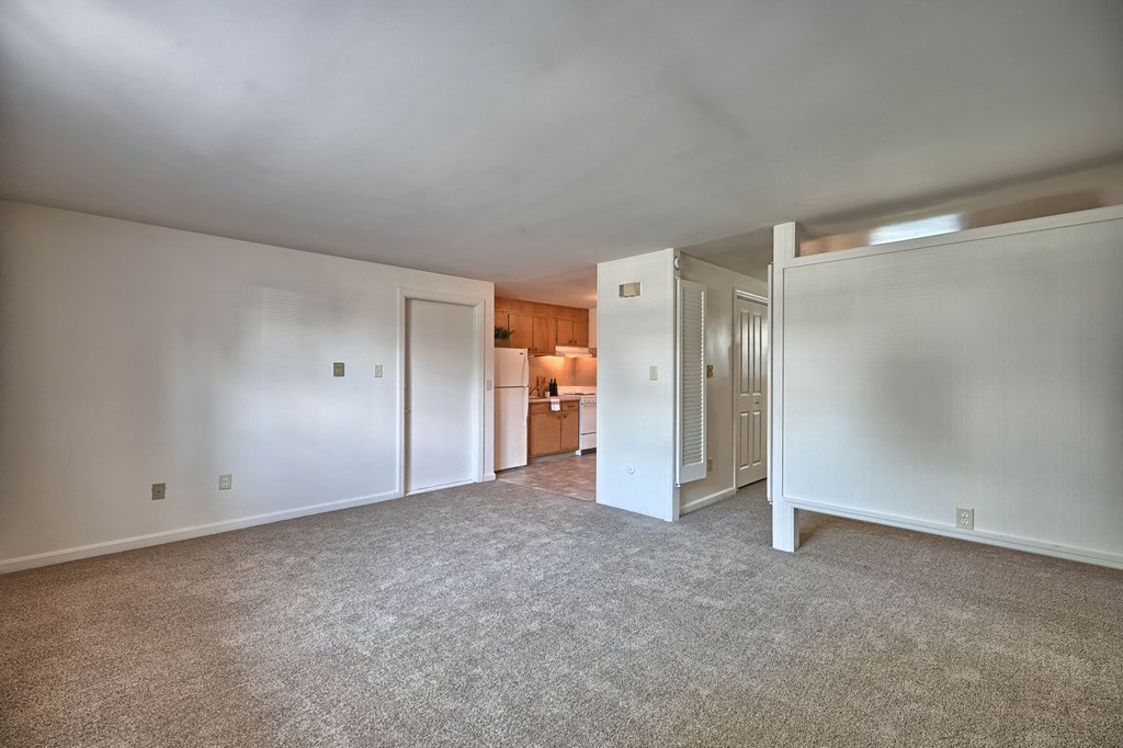a living room with white walls and carpet and a kitchen