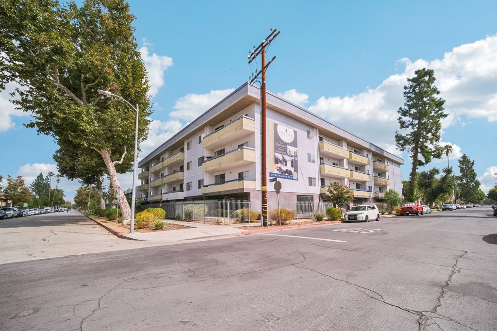A white building with a blue sign on the front is surrounded by trees and a street.