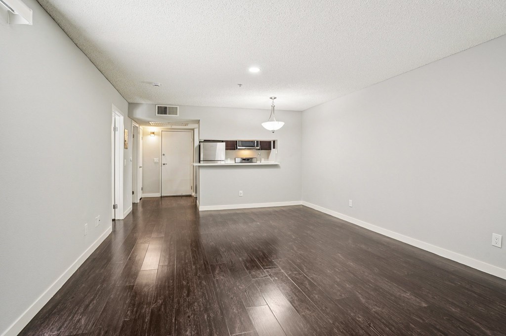 A room with wooden floors and a kitchen area in the background.