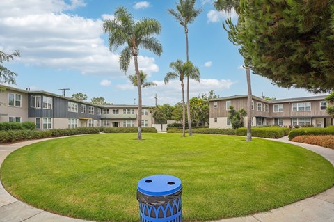 A blue trash can sits in the middle of a grassy circle in front of apartment buildings.