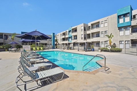 A swimming pool surrounded by sun loungers in front of apartment buildings.