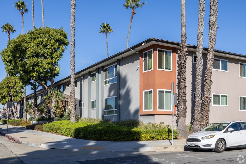 A white car is parked in front of a two-story building with a red stripe on the side.