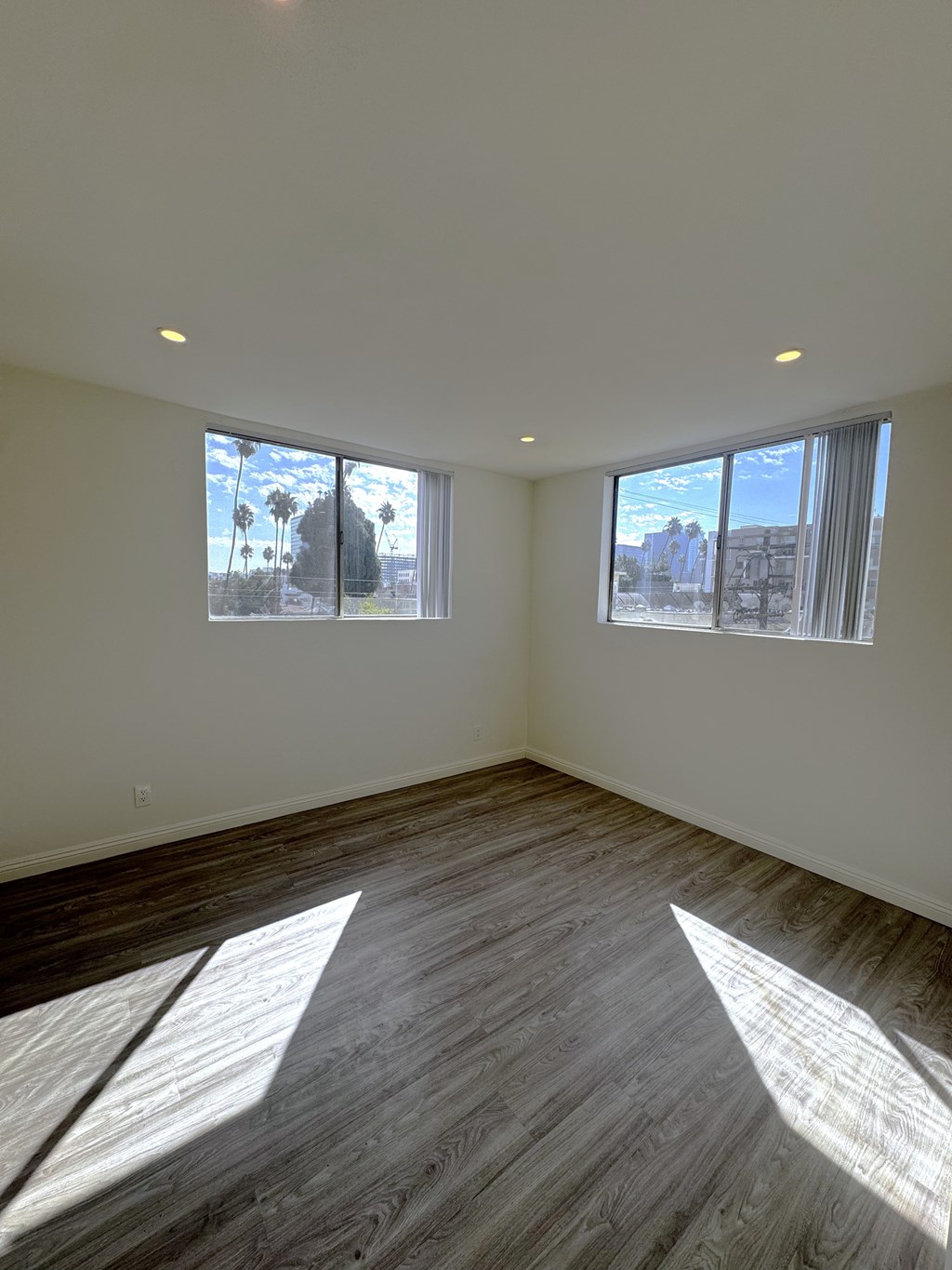 A room with wooden flooring and two windows showing a view of a street and buildings outside.