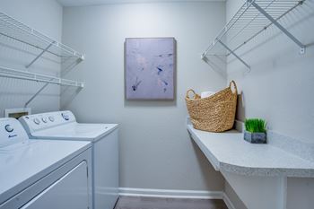 a laundry room with a washer and dryer and a shelf with a basket at The Monroe Apartment Homes, Tallahassee, 32303