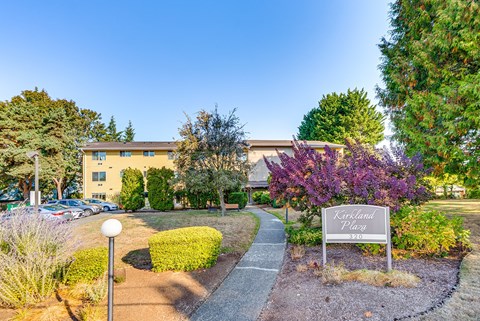 The image shows a sign that reads "Kirkland Place" in front of a building with a parking lot and trees in the background.