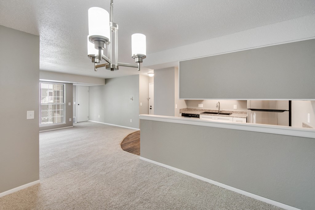 A well-lit, empty room with a large window and a kitchen area in the background.