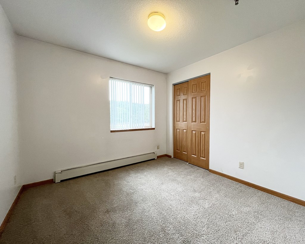 A room with a brown carpet, a window with white blinds, a wooden door, and a white wall.