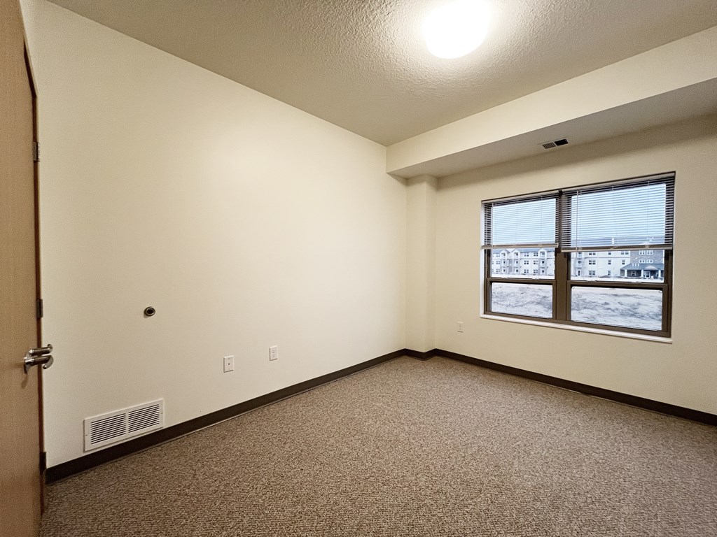 A room with a carpeted floor, a window with a view of other buildings, and a door on the left side.