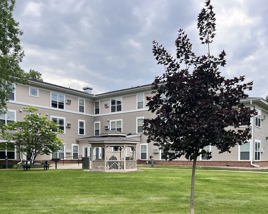 A tree stands in front of a building with a gazebo in the middle of a grassy courtyard.