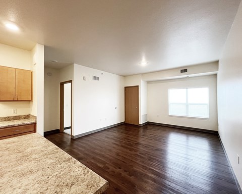 A large empty room with wooden floors and a kitchen area to the left.