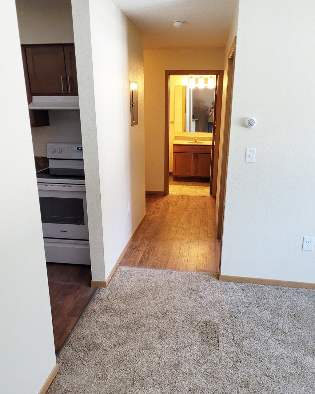 an empty room with a carpeted floor and a hallway to a kitchen and a