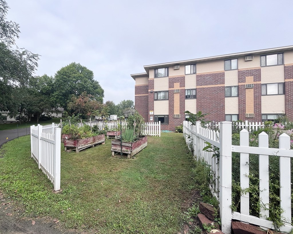 A white picket fence surrounds a small garden in front of a brick apartment building.