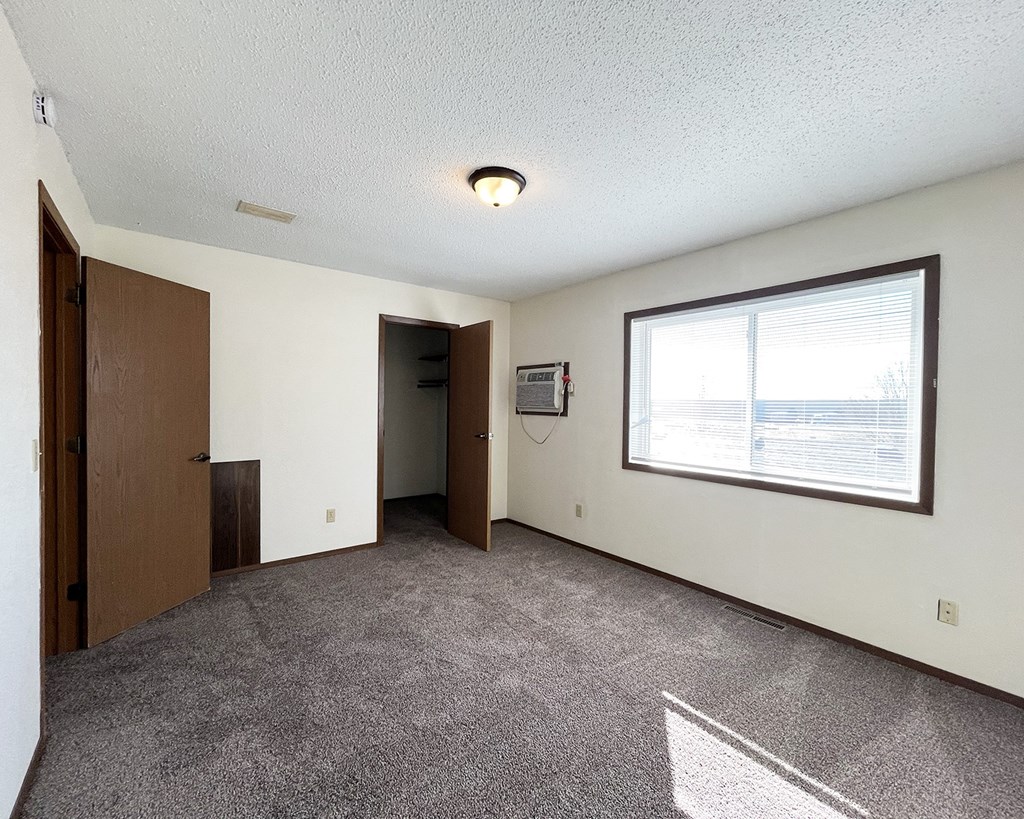 A room with a carpeted floor, a window with blinds, and a door leading to another room.