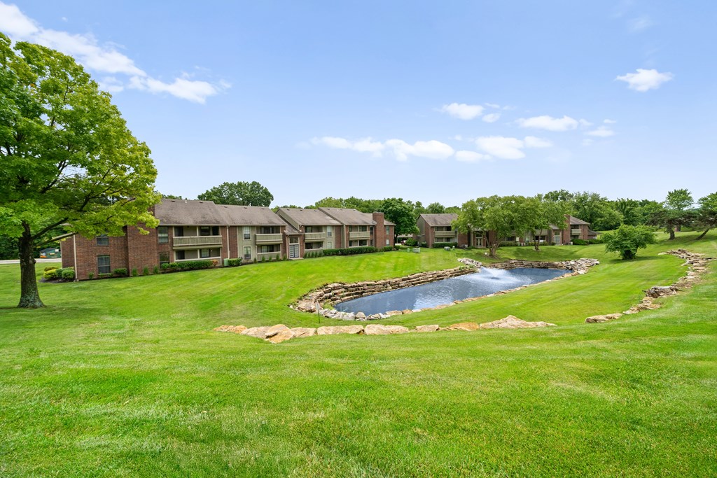 A small pond in the middle of a grassy area with a building in the background. at Coventry Oaks Apartments, Overland Park, KS, 66214