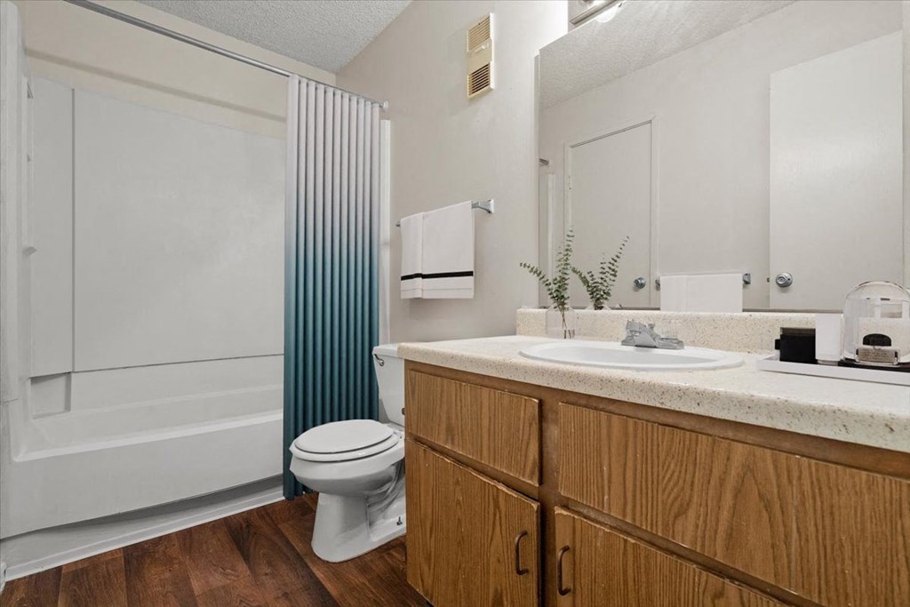 A bathroom with wooden storage cabinets, a large vanity mirror, lots of counter space, a toilet, and a tub shower combo at Bristol Pointe Apartment Homes, Olathe, Kansas