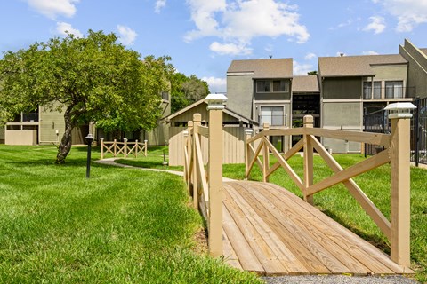 A wooden bridge crosses a grassy area in front of apartment buildings.