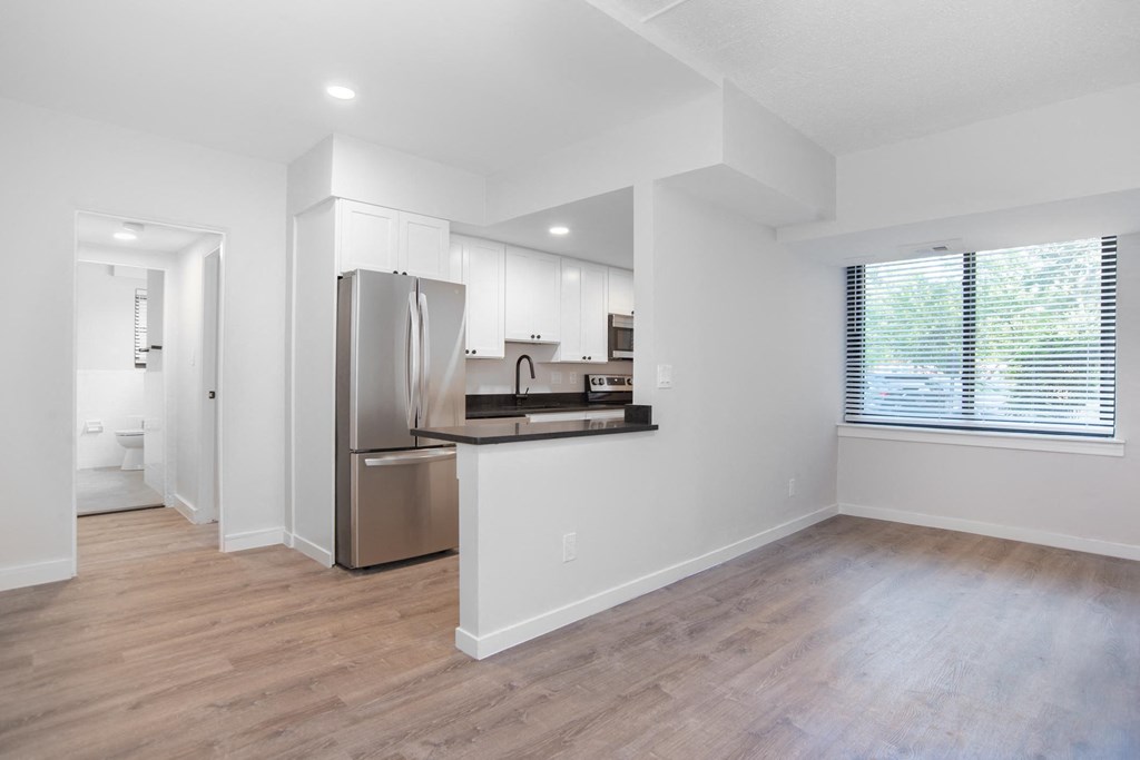 an empty living room with a kitchen with a stainless steel refrigerator