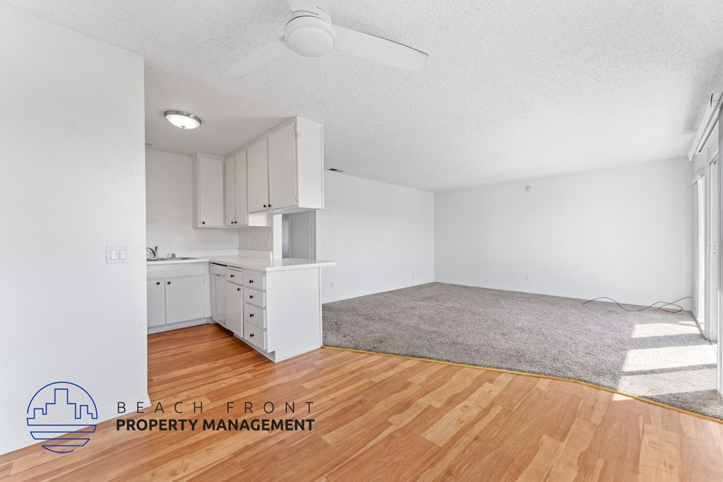 A room with wooden floors and white walls, featuring a kitchen area with white cabinets and a countertop.