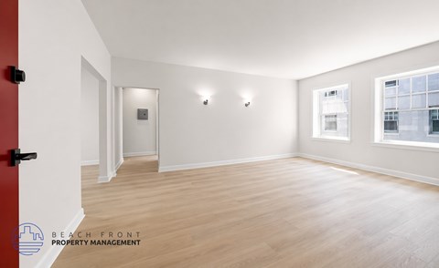 A spacious room with wooden flooring and white walls, featuring a red door and a window with a view of a building outside.