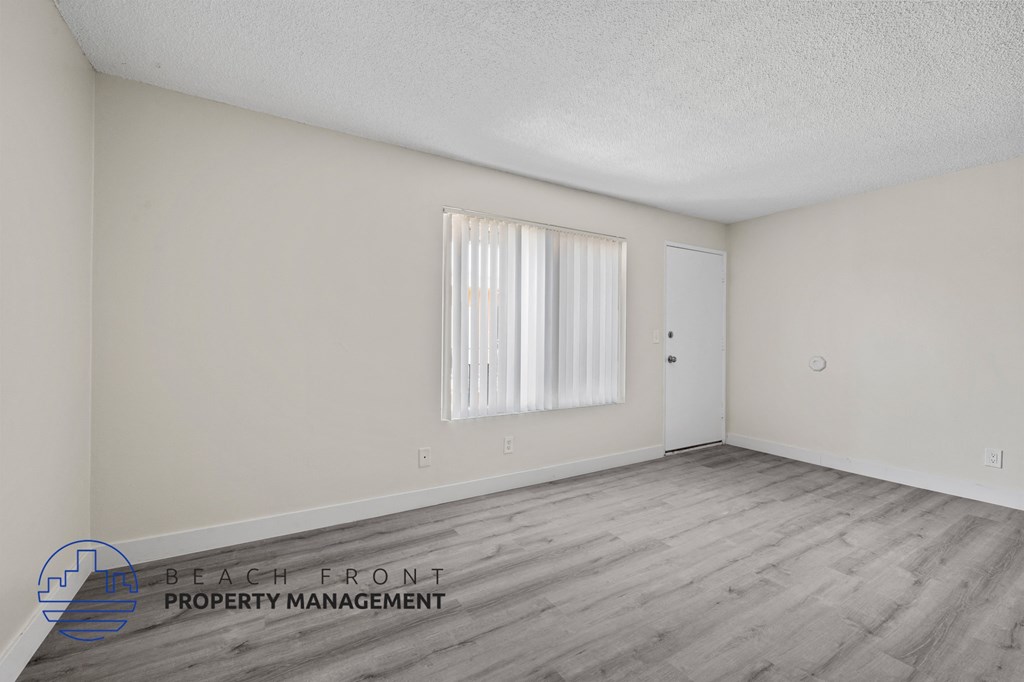 A spacious room with wooden flooring and a window with white curtains.