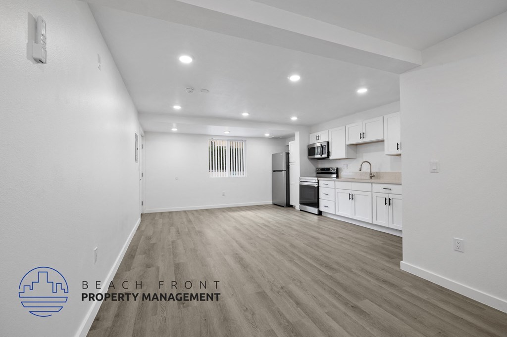 A spacious, empty room with a kitchen area in the background and a logo for Beach Front Property Management in the bottom left corner.