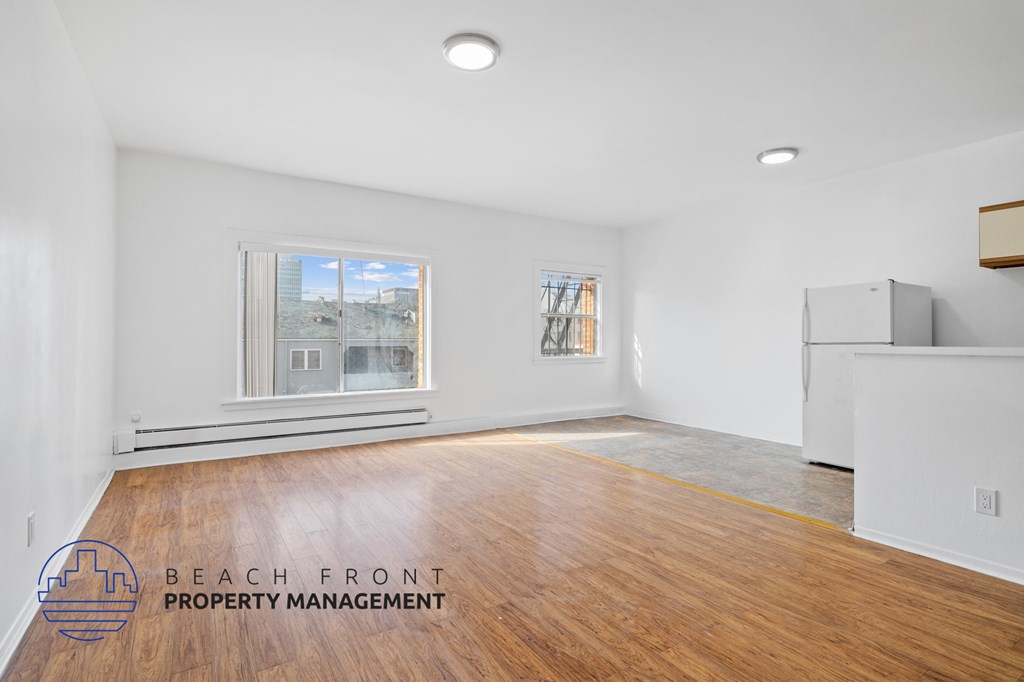 A spacious room with wooden flooring and a large window overlooking a beachfront.
