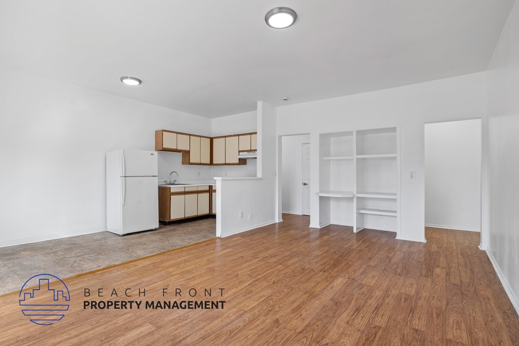 A spacious room with wooden floors and white walls, featuring a kitchenette with a refrigerator, sink, and cabinets.