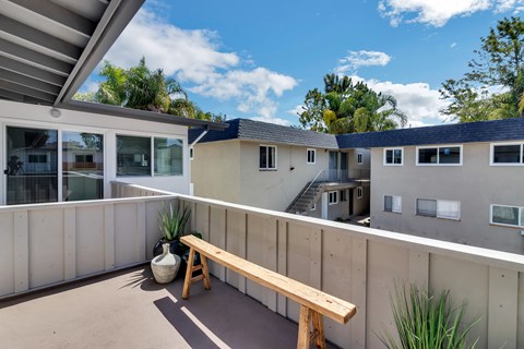 A wooden bench sits on a patio with a concrete wall and a building in the background.
