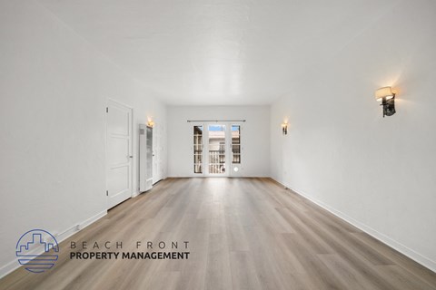 A spacious room with wooden floors and white walls, with a logo for Beach Front Property Management in the bottom left corner.