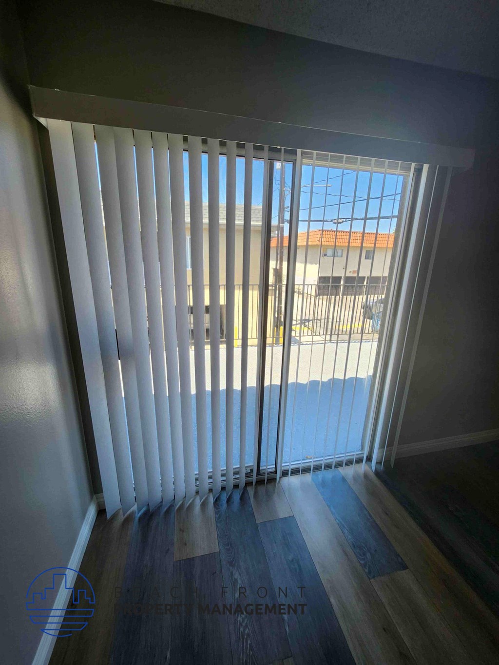 A room with a view of a building through a window with blinds.