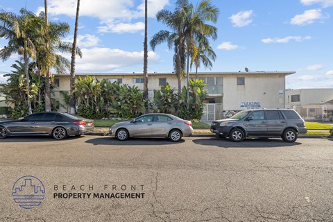 Three cars are parked in a lot with a building and palm trees in the background.