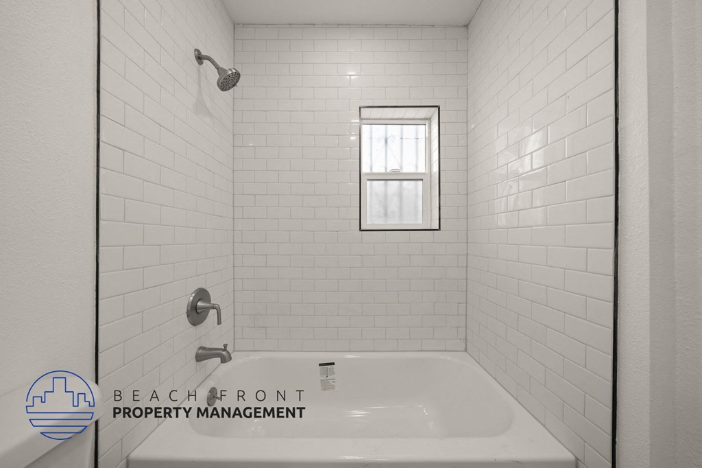 A white tiled bathroom with a shower, sink, and window.