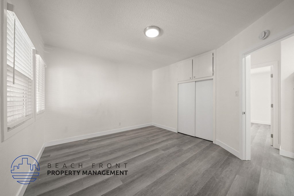 A spacious room with wooden flooring and white walls, with a logo for Beach Front Property Management in the bottom left corner.