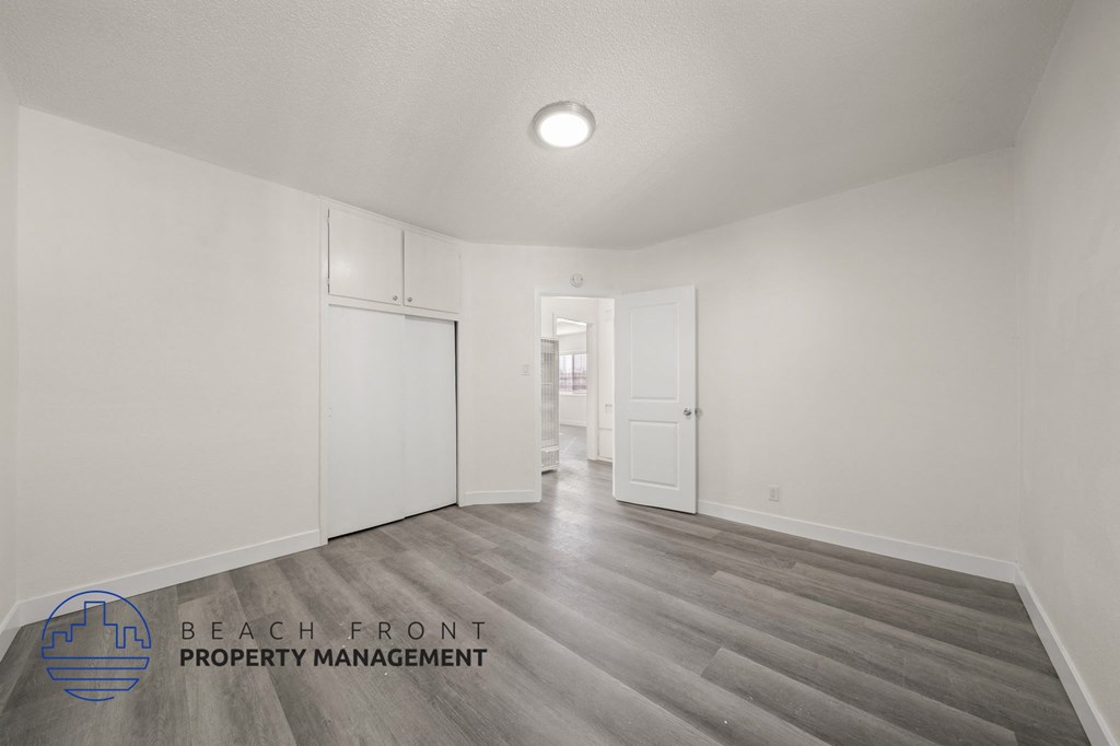A spacious room with wooden flooring and white walls, with a logo for Beach Front Property Management in the bottom left corner.